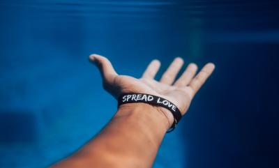 human reaching out their hand, with a wrist band with writing on it saying "Spread Love"