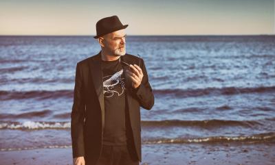 A man walking in a dark jacket, t-shirt and hat, by the sea