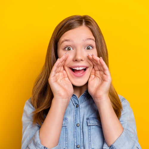 Girl speaking into her hands, she's stood in front of a bright yellow wall.